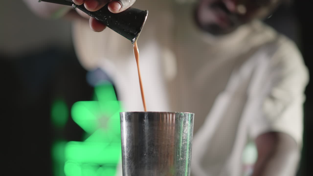Bartender pouring liquid from jigger into cocktail shaker at bar counter with neon green lighting, focused on precise drink preparation in vibrant bar atmosphere