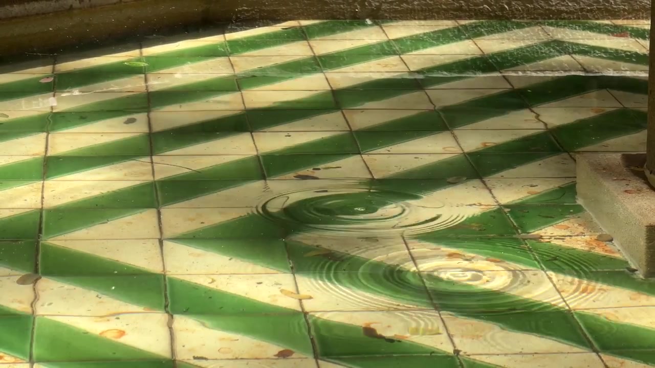 Statisk 4K nærbillede af grønne og hvide fliser på springvandsbunden under lavt vand. Blide dråber skaber bløde krusninger på overfladen i skarpt dagslys