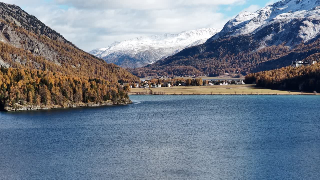 Drone flying forward over alpine lake with snowy peaks and autumn trees in Switzerland's Engadin valley