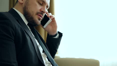 close-up tilt down of confident businessman talking mobile phone and writing notes in notepad while sits on armchair in hotel room or office. travel, business and people concept