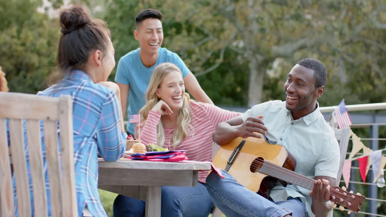 African american man playing guitar with diverse friends at dinner table in garden, slow motion