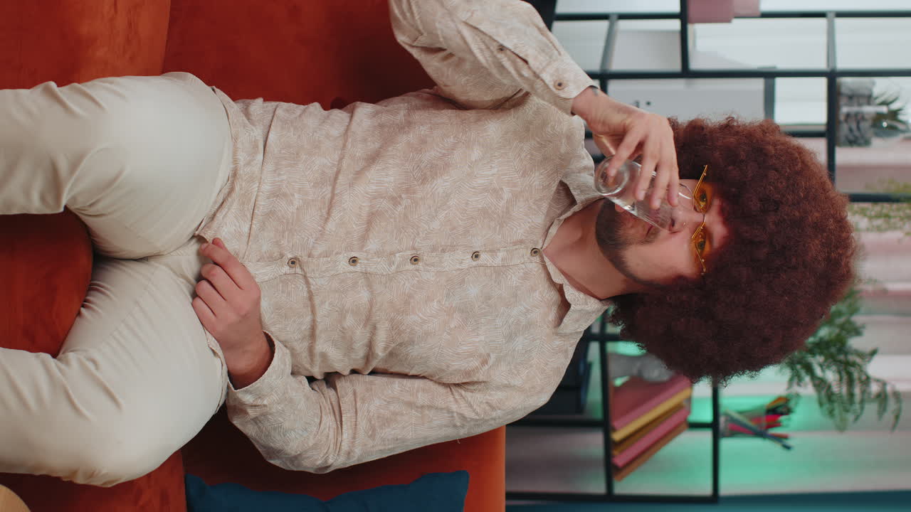 Thirsty young adult man sitting at home holding glass of natural aqua make sips drinking still water
