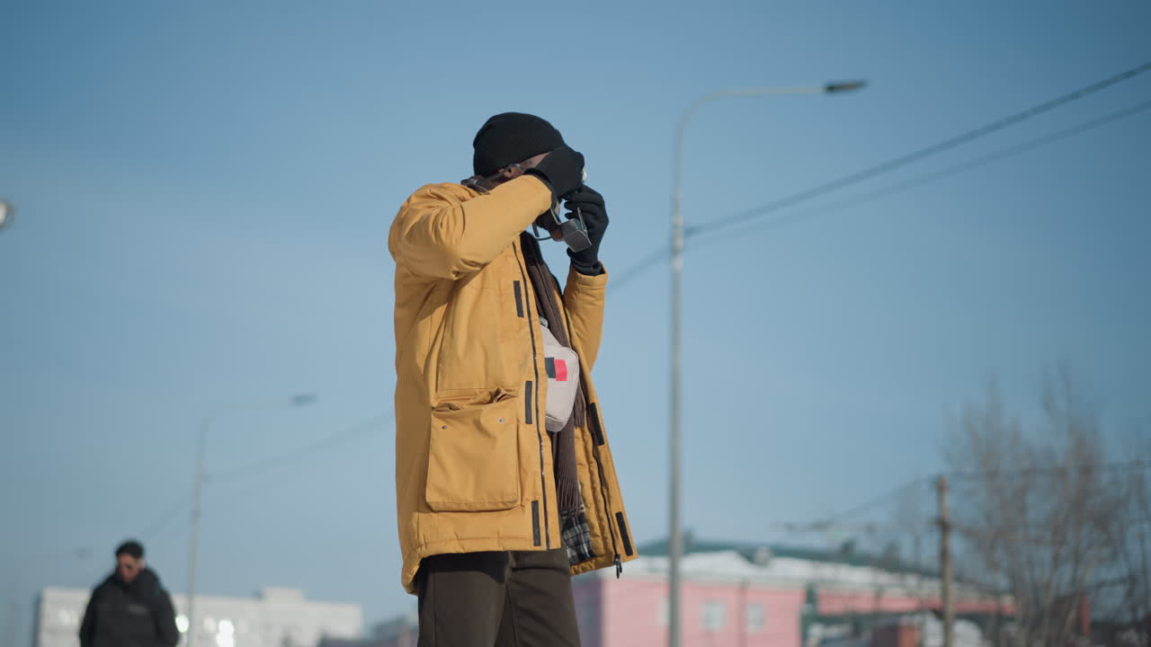 cameraman in yellow coat gloves black beanie and scarf aiming vintage camera up adjusting focus through viewfinder on snowy city street under bright winter sun with pedestrians walking in background