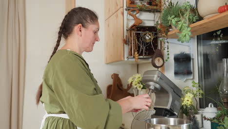 old woman in green dress and white apron fixes whisk attachment into electric mixer inside cozy kitchen with wooden cabinets and plants, preparing for mixing ingredients in stainless bowl