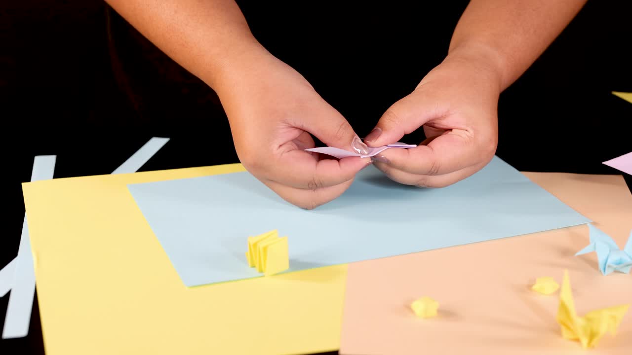Person folds pastel origami paper on colored sheets under bright lighting, close-up overhead view