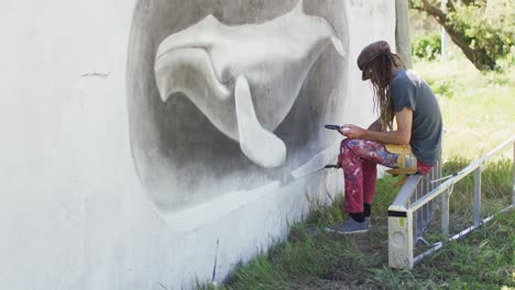 video de un artista masculino caucásico con rastas sentado en una escalera pintando un mural de ballena en la pared