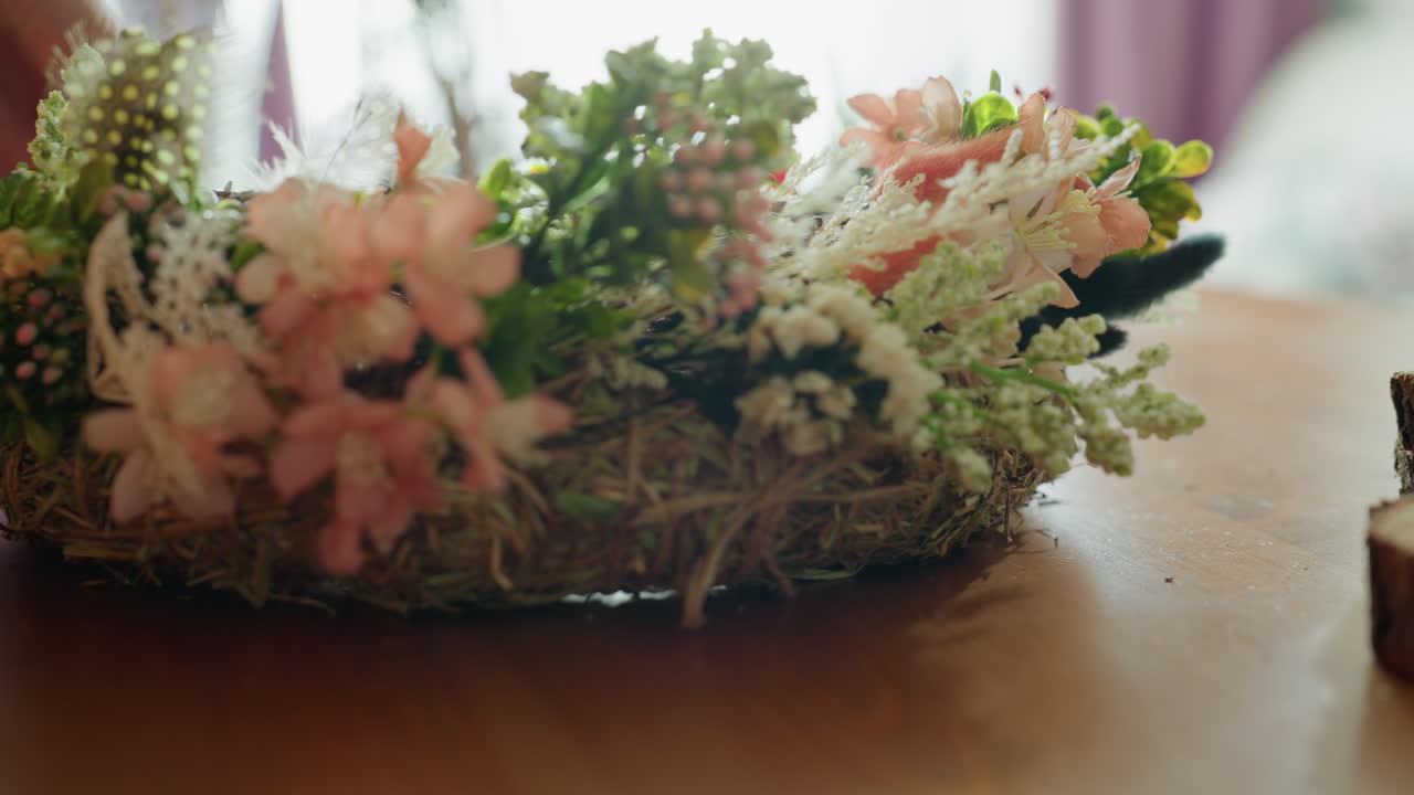 Hand arranging rustic floral wreath with twigs, hay, and pink blossoms on wooden table, surrounded by moss pieces and natural decorations, artisan crafting handmade decorative centerpiece