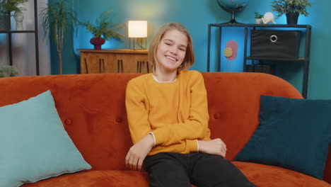 teenager boy waving with smile greeting happily sitting on sofa looking straight at camera at home