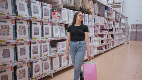 lady in black top and jeans walking through supermarket aisle holding shopping bags, focusing on products displayed on shelves, neatly arranged bedding and home textiles in bright retail environment