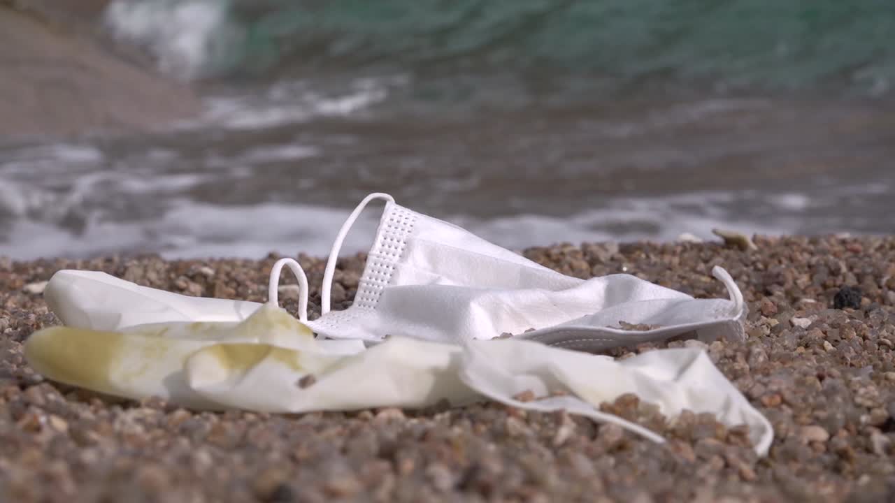 해변에서 버려진 마스크와 장갑, 근처에 연속적인 돌진 파도가 있고, 꾸준한 샷이 왼쪽으로 이동합니다.