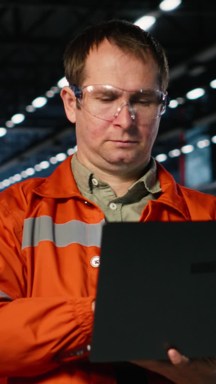 Vertical Video Specialist in protective uniform observes logistics via laptop on factory floor
