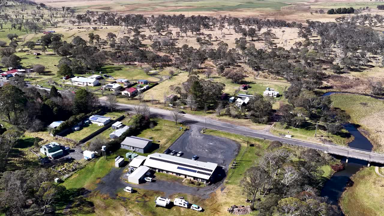 Drone footage glides above a small rural town with scattered houses, roads, and a creek, under bright daylight, revealing the surrounding countryside landscape
