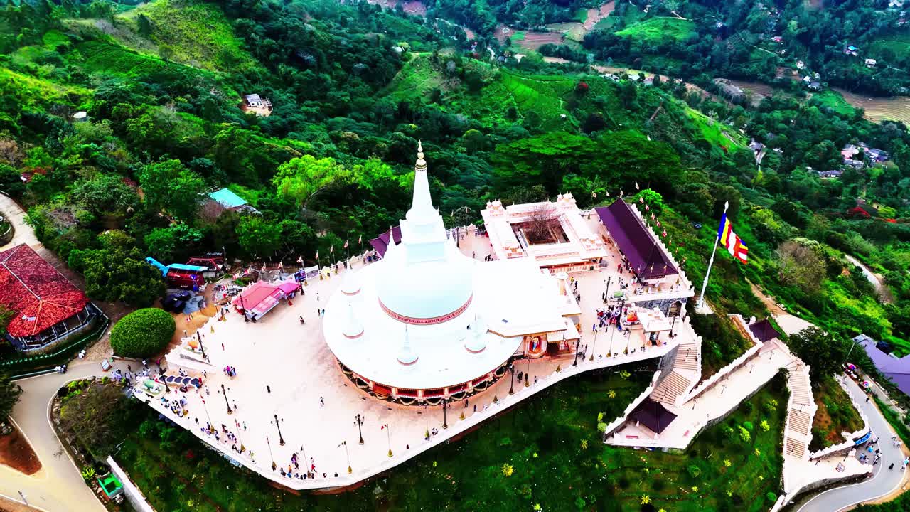 Striking aerial shot of a large white Buddhist stupa and temple complex in Sri Lanka, bustling with visitors, set atop lush green hills with winding paths and traditional buildings nearby.