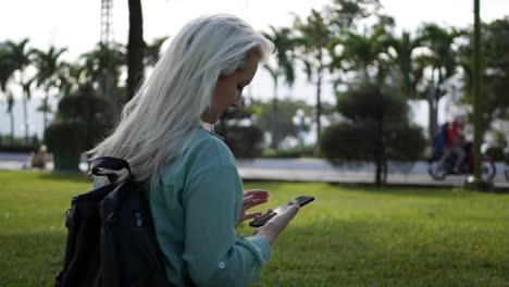 hermosa mujer delgada con el cabello largo rubio en camisa verde se sienta en el suelo y el uso de teléfono inteligente sobre el fondo del parque. niña en la pantalla cuadrada tocando y sonriendo