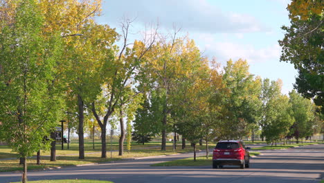 Red-car-driving-away-from-camera-in-the-fall-in-Calgary,-Alberta,-Canada-in-a-nice-neighbourhood