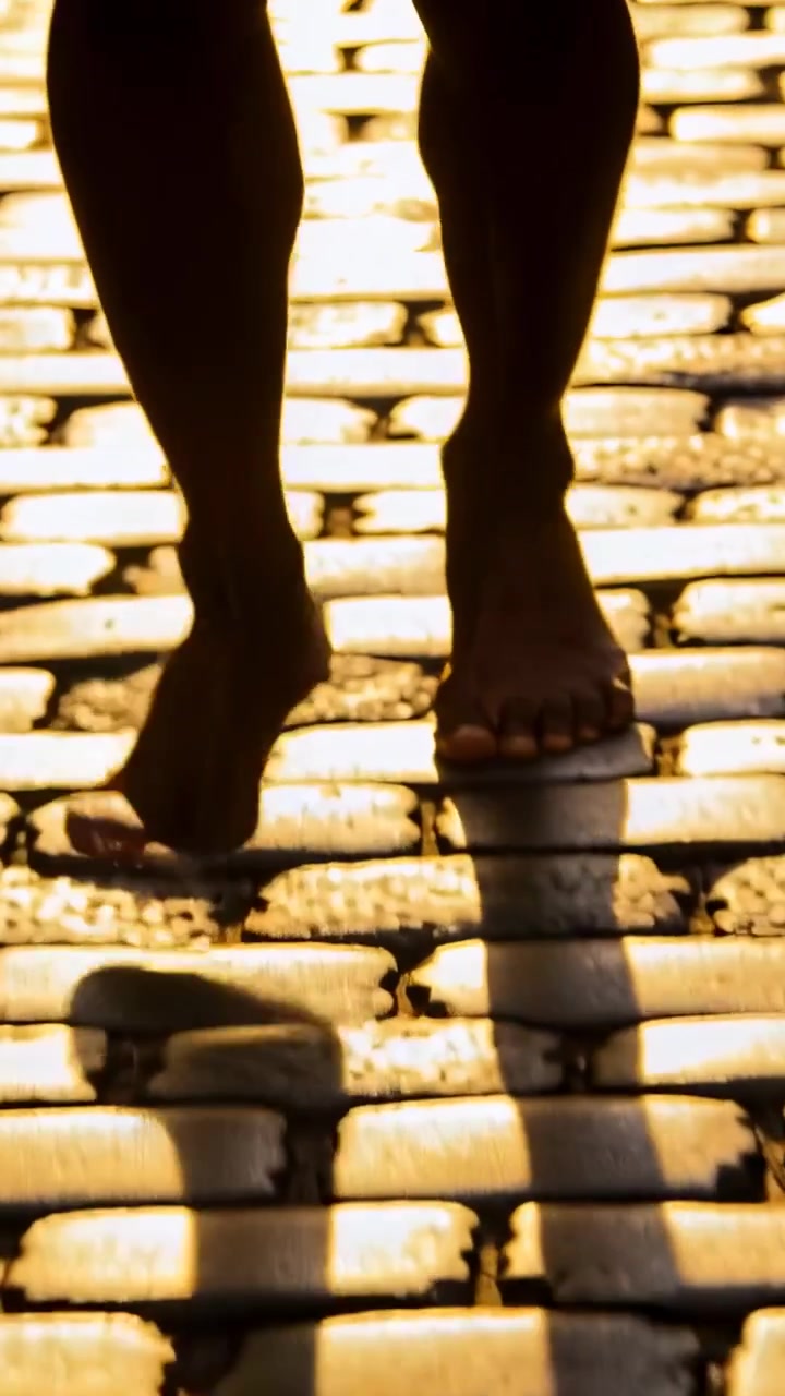 Video verticale: Un adulto a piedi nudi che solleva il piede e cammina verso la telecamera su un selciato, proiettando lunghe ombre.