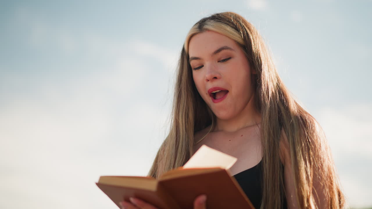 primer plano de una joven pensativa vestida de negro hojeando un libro al aire libre bajo un cielo brillante, encontrando un papel escondido dentro de las páginas y sonriendo emocionada, mientras la luz del sol ilumina suavemente su rostro