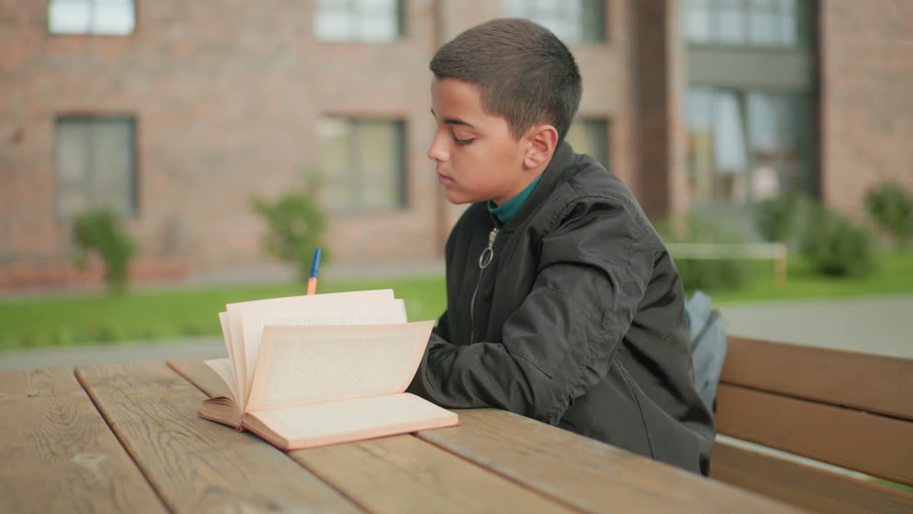 Side view of kid seated outdoors writing in notebook with open book as pages flip in wind, soft blur background showing greenery and building details, symbolizing focus during study