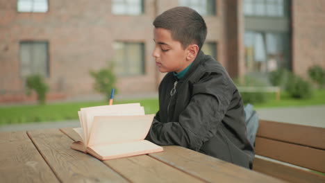 Side view of kid seated outdoors writing in notebook with open book as pages flip in wind, soft blur background showing greenery and building details, symbolizing focus during study