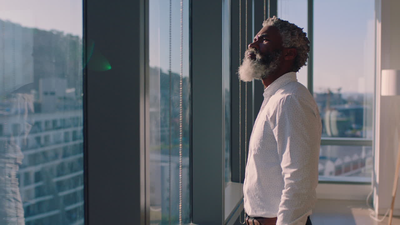 hombre de negocios afroamericano maduro mirando por la ventana planeando hacia adelante pensando en ideas para el desarrollo de negocios futuros con vista a la ciudad al atardecer 4k