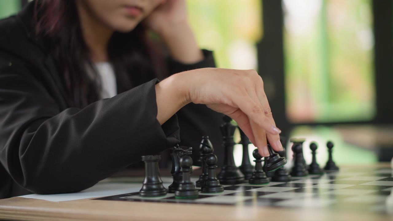 Close-up de jovem mulher jogando xadrez, movendo peças pretas no tabuleiro. A cena mostra sua expressão focada e movimentos das mãos com componentes de jogo de tamanho padrão em luz natural suave.