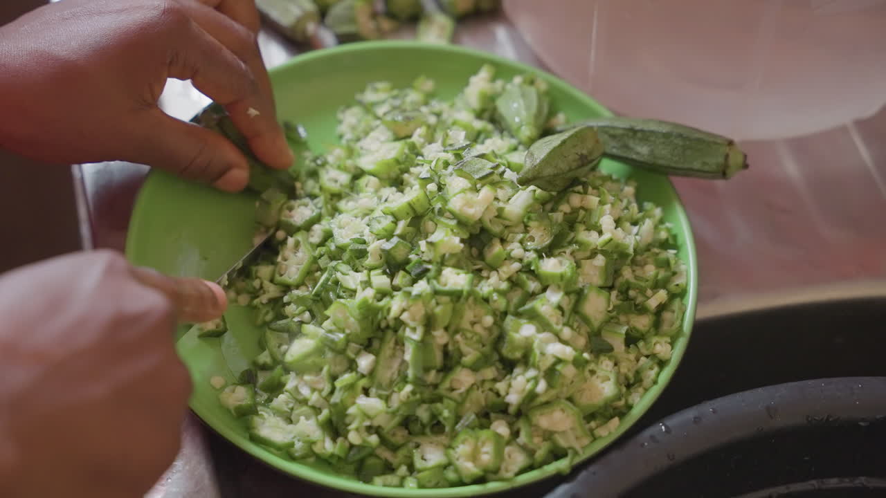 Okra picada en un cuenco verde, manos negras moviéndose, preparación al estilo de vendedor con rebanado y enjuague, manos revolviendo y ajustando el condimento, primer plano de textura y movimiento, energía vibrante de cocina de mercado.
