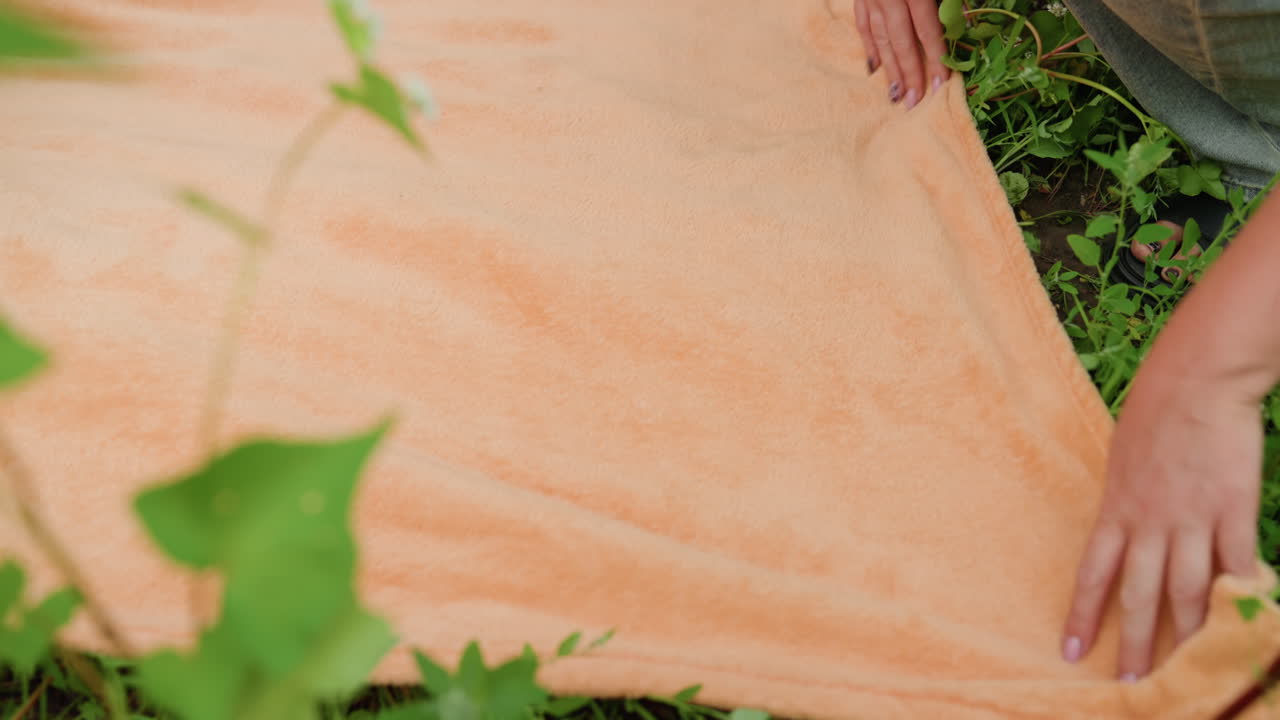 Primer plano de una mujer blanca alisando una toalla naranja en el exterior con movimientos suaves mientras extiende la tela sobre un prado verde con flores silvestres; se aprecia la textura agradable del tejido.