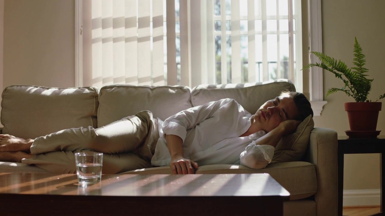 Streaming sunlight lulling man in white shirt into rest on sofa at home, glass of water