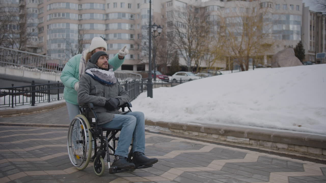 Happy Caucasian woman taking her disabled friend in wheelchair for a walk in the city and talking together