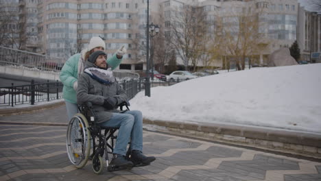 happy caucasian woman taking her disabled friend in wheelchair for a walk in the city and talking together