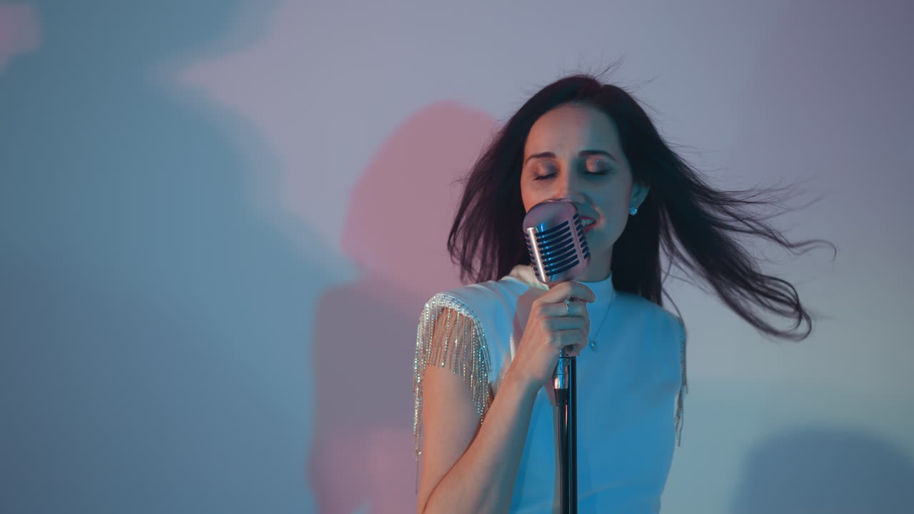 A singer in a white dress passionately sings, holding a vintage microphone with one hand against a blue background. Her hair flows back