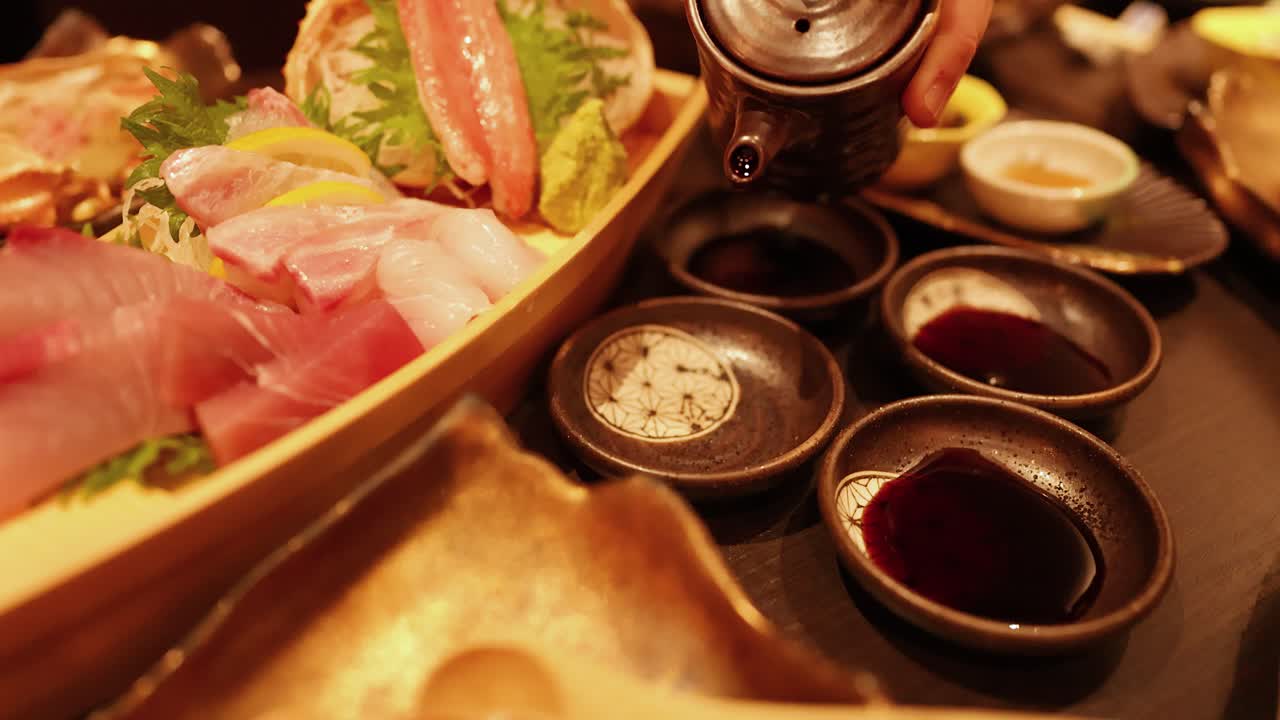 Una mano versa salsa di soia in ciotole di ceramica accanto a una barca di legno con sashimi fresco.