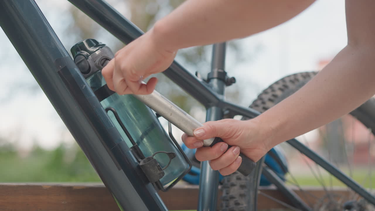 Woman removes pump from bike frame. Closeup of hands unlocking metal bracket and detaching compact pump from frame near rear tire, park bench background, morning light, focused practical mood