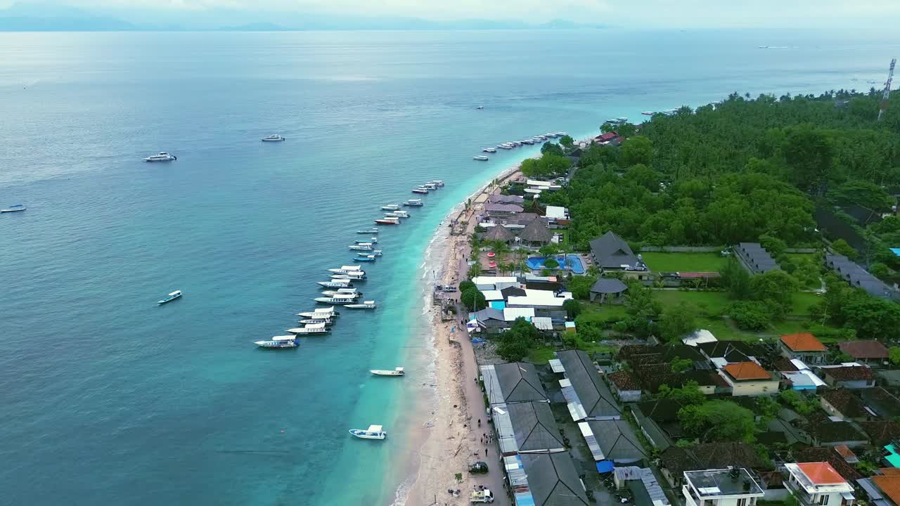 Drone view shows Padangbai coastline with clear turquoise water, long sandy beach, anchored boats, green hills, and coastal village creating calm tropical atmosphere in eastern Bali