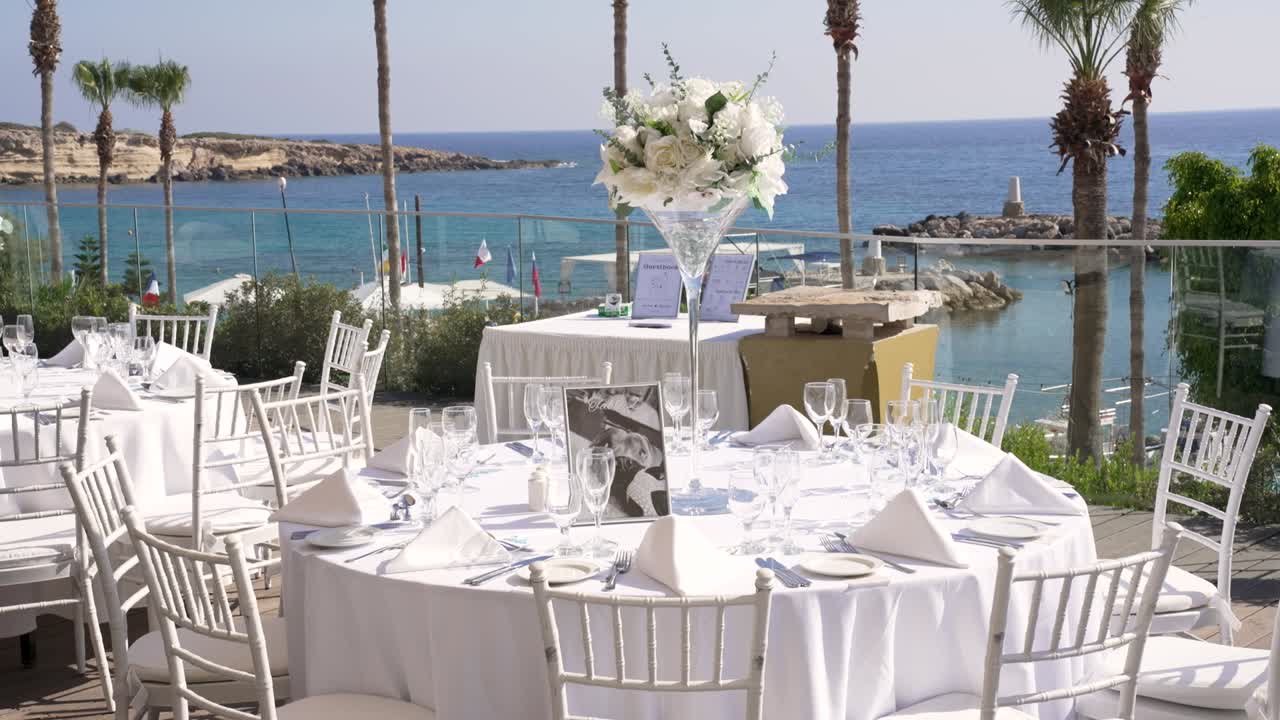 Elegant tables are arranged by the sea for a memorable gathering. White linens and floral centerpieces create a stunning backdrop against the ocean view