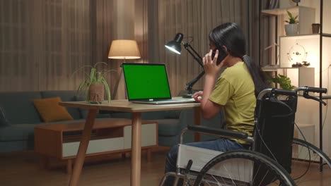 back view of asian kid girl sitting in a wheelchair while using laptop computer with mock up green screen and talking on mobile phone at home