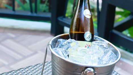 a wine bottle rests in an ice bucket on a patio, surrounded by greenery, under bright daylight