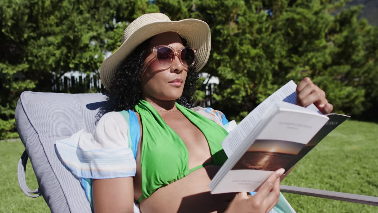 African American woman lounging on lawn chair wearing hat, reaching, turning book pages to relax
