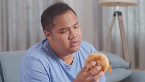 close up of a fat asian man shaking his head being bored having fried chicken while eating fast food on a sofa in the living room at home