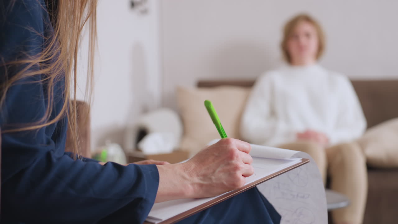 Woman seated on sofa expresses emotions during session with supportive listener who attentively takes notes, showcasing therapeutic conversation, trust, and emotional openness in calm indoor setting