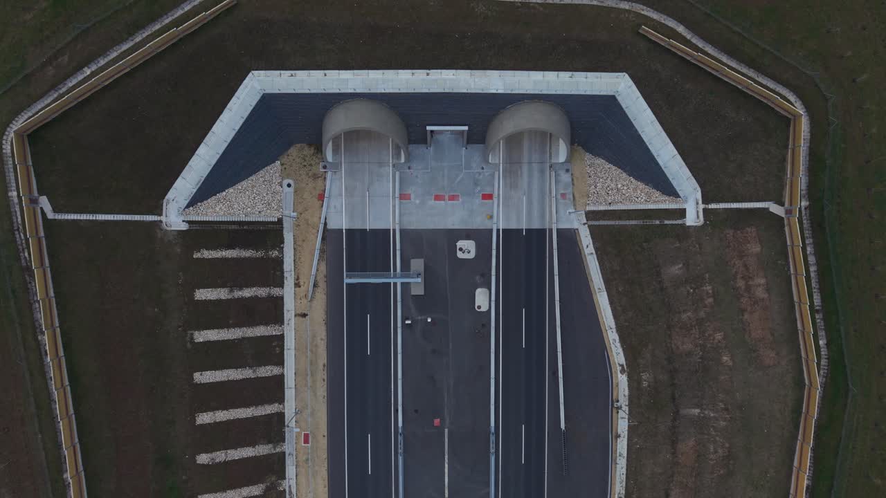 A top-down aerial view of a dual highway tunnel entrance featuring clean concrete structures and modern road design. Vehicles are visible entering and leaving the tunnels