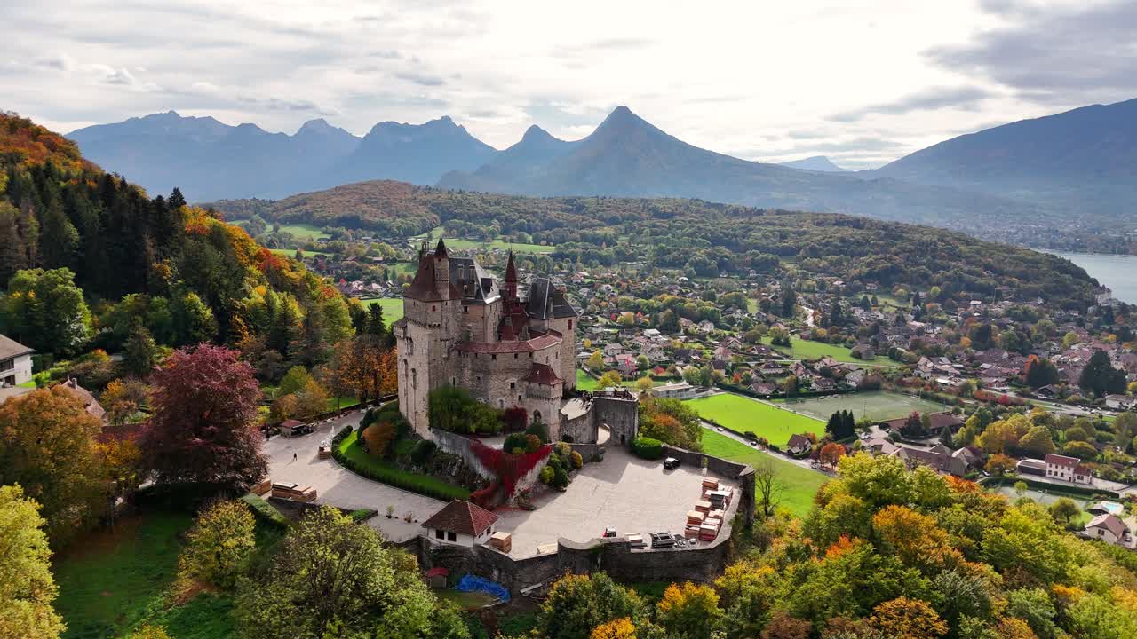 Aerial view of Burg Menthon-Saint-Bernard in France surrounded by colorful autumn forests, nearby village homes, open fields and distant mountain peaks under a softly clouded sky