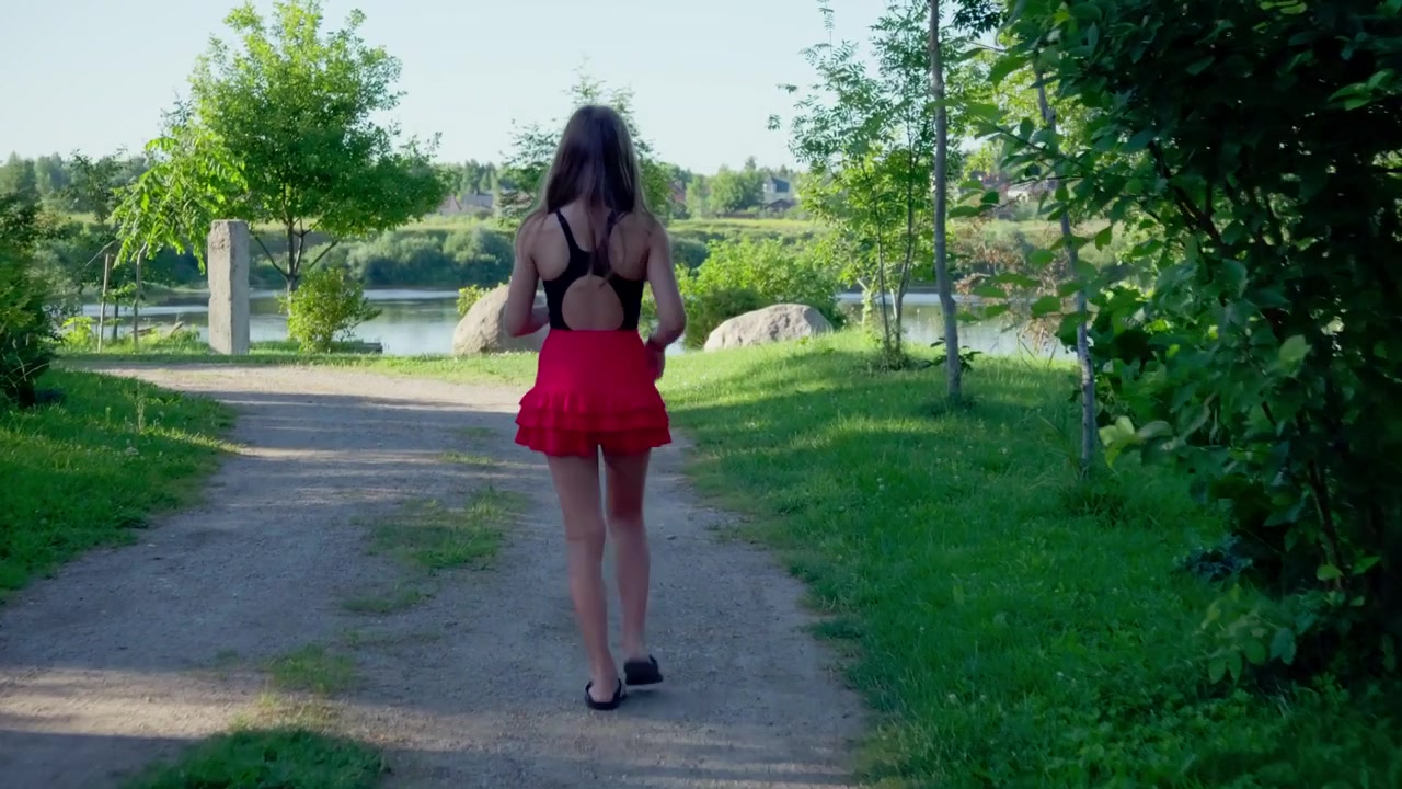 Ripresa con telecamera a seguire di una giovane ragazza con una gonna rosso brillante che passeggia su un sentiero sterrato attraverso un lussureggiante parco verde verso un fiume panoramico, catturando un sereno ambiente estivo all'aperto.