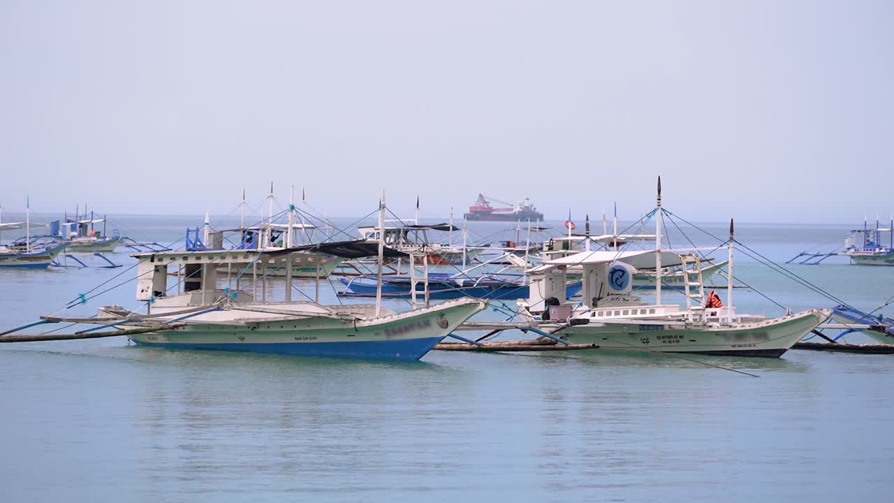 穏やかな熱帯の海に停泊する伝統的なアウトリガー漁船。地平線には遠くの貨物船が見え、地元の漁業と商業海運活動の対比を示しています。