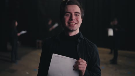 Portrait-of-a-confident-and-happy-brunette-male-actor-in-a-black-suit-who-reads-his-text-and-then-folds-piles-of-papers-near-his-chest-near-the-actors-who-are-preparing-to-perform-on-stage-in-the-theater