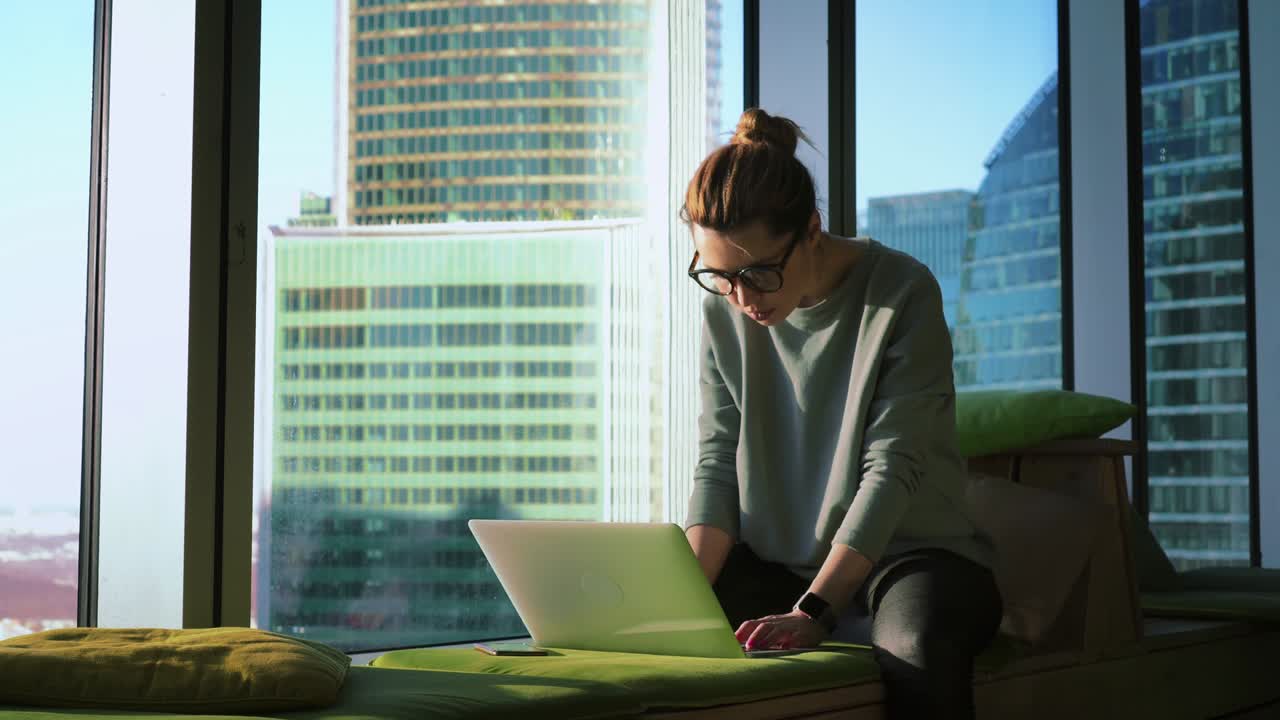 Donna d'affari con gli occhiali è seduta su un divano morbido vicino a una finestra panoramica e sta aprendo il laptop per iniziare a lavorare