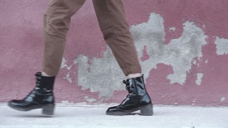 Slow-motion-close-up-of-a-woman-walking-forward-wearing-shiny-black-boots-and-brown-pants-at-an-urban-set-up-with-a-red-wall-as-a-background