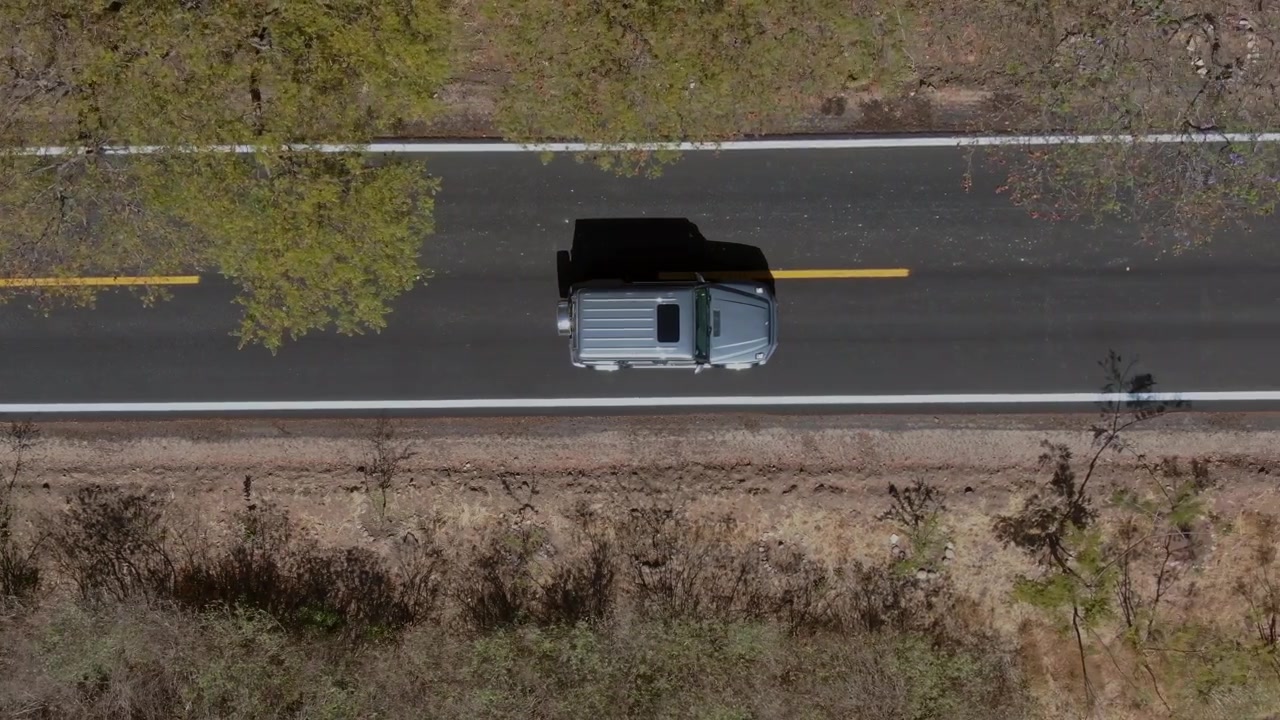 Vista aérea con dron de una camioneta circulando por una carretera rural a través de un paisaje árido. Perspectiva cenital que muestra el vehículo, la carretera, la vegetación de la cuneta y el terreno seco.