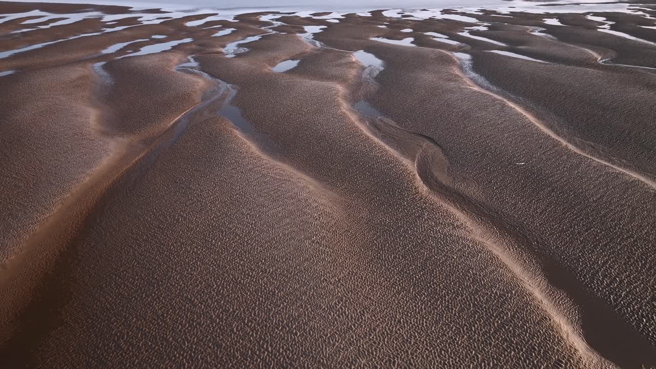 Lateral flight over undulating sand patterns with seawater pools at sunset. Aerial footage.