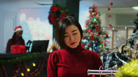 portrait of happy asian woman in red sweater browsing through clothes racks in festive shopping mall clothing store during christmas holiday season. shopper looking to buy gifts in fashion boutique